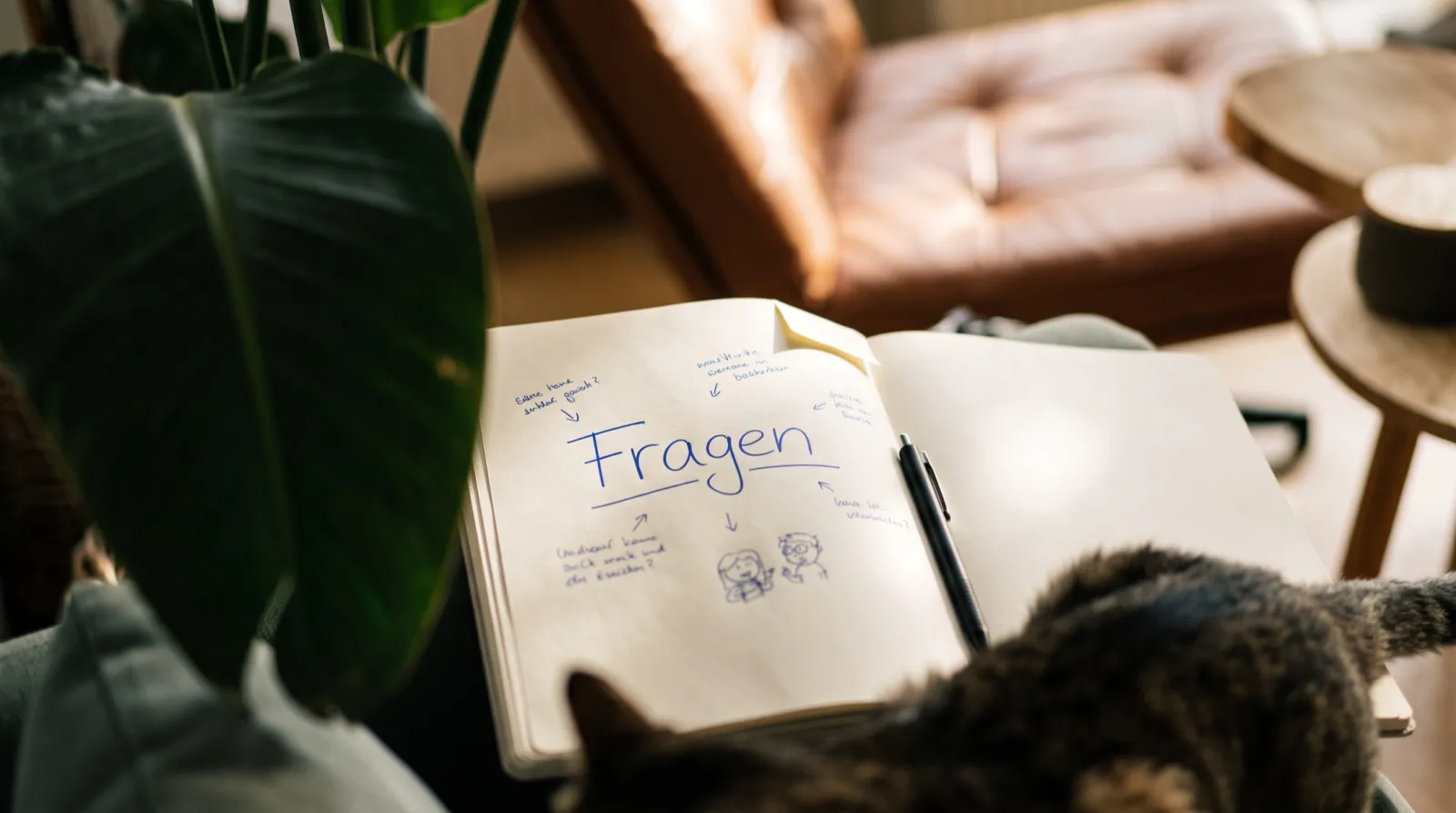 Ein aufgeschlagenes Notizbuch auf einem Sofa vor einem Ledersessel, darauf das Wort Fragen in blauer Handschrift.